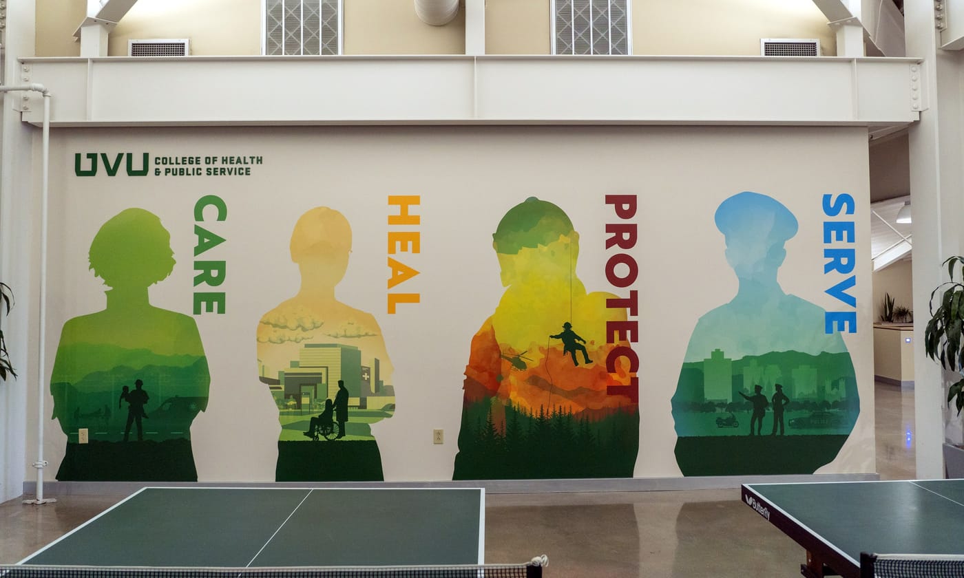 A long wall in a college commons printed with four tall silhouettes labeled Care, Heal, Protect and Serve, each filled with scenes of health and public-service work.