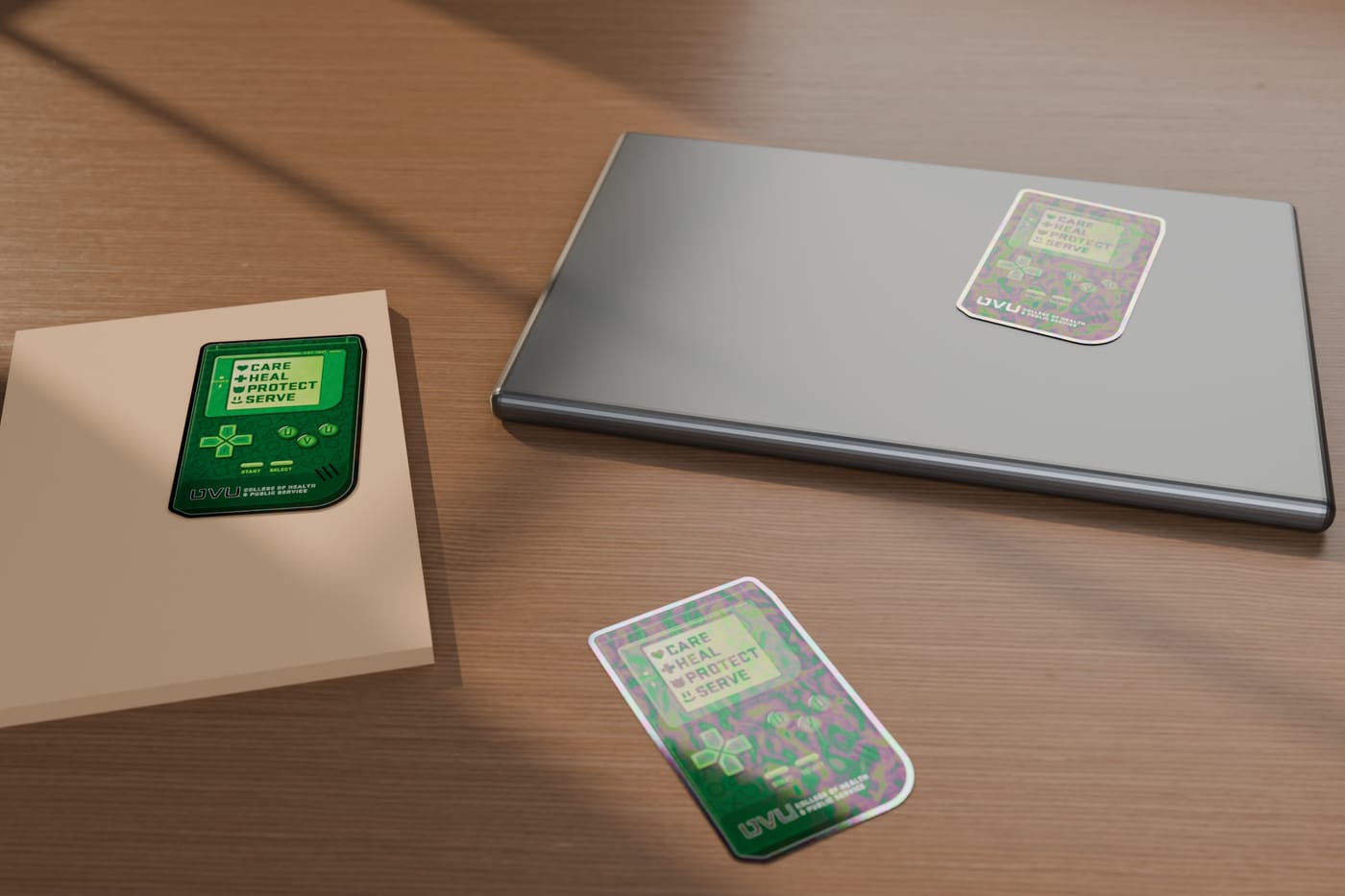 Holographic stickers of a green handheld game console listing Care, Heal, Protect, Serve, on a laptop lid, a kraft notebook and a wooden desk in window light.