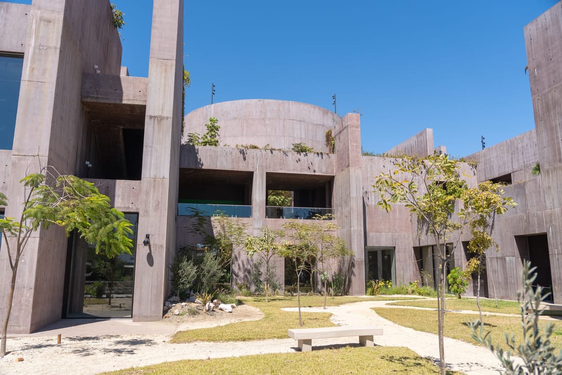 Courtyard of pinkish board-formed concrete with a domed volume, young trees, a bench and gravel paths under a blue sky.