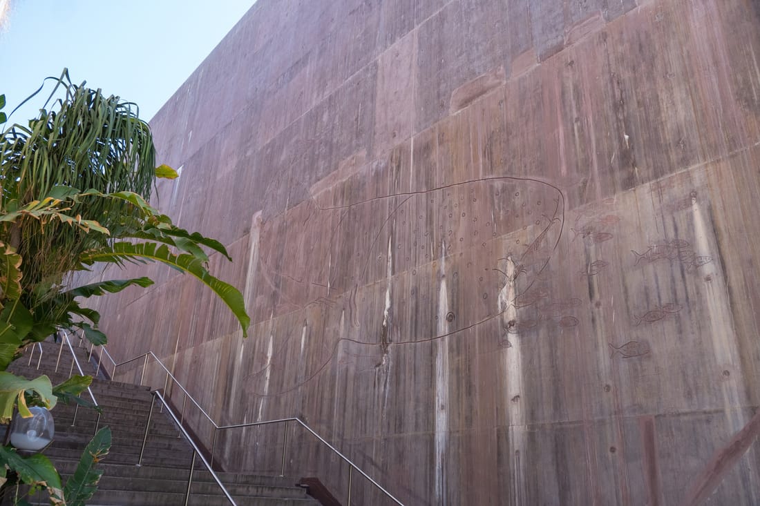 A towering board-formed concrete wall beside an outdoor stair, with a tropical plant in the foreground.