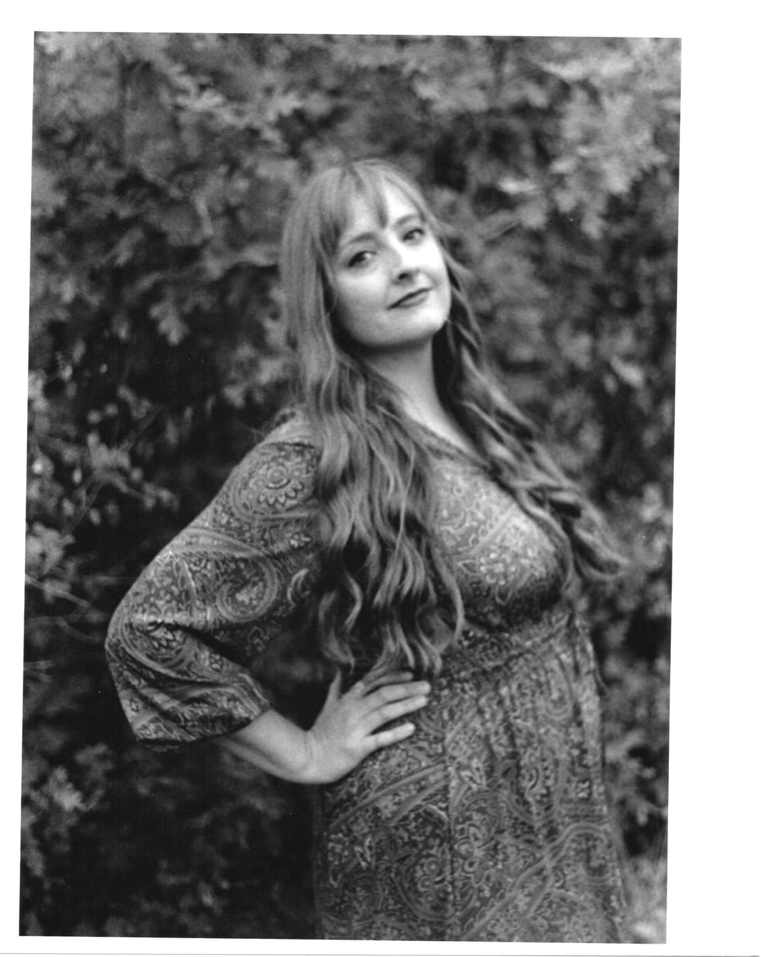 Print of a woman with long wavy hair and a patterned dress, hand on hip, in front of leaves.
