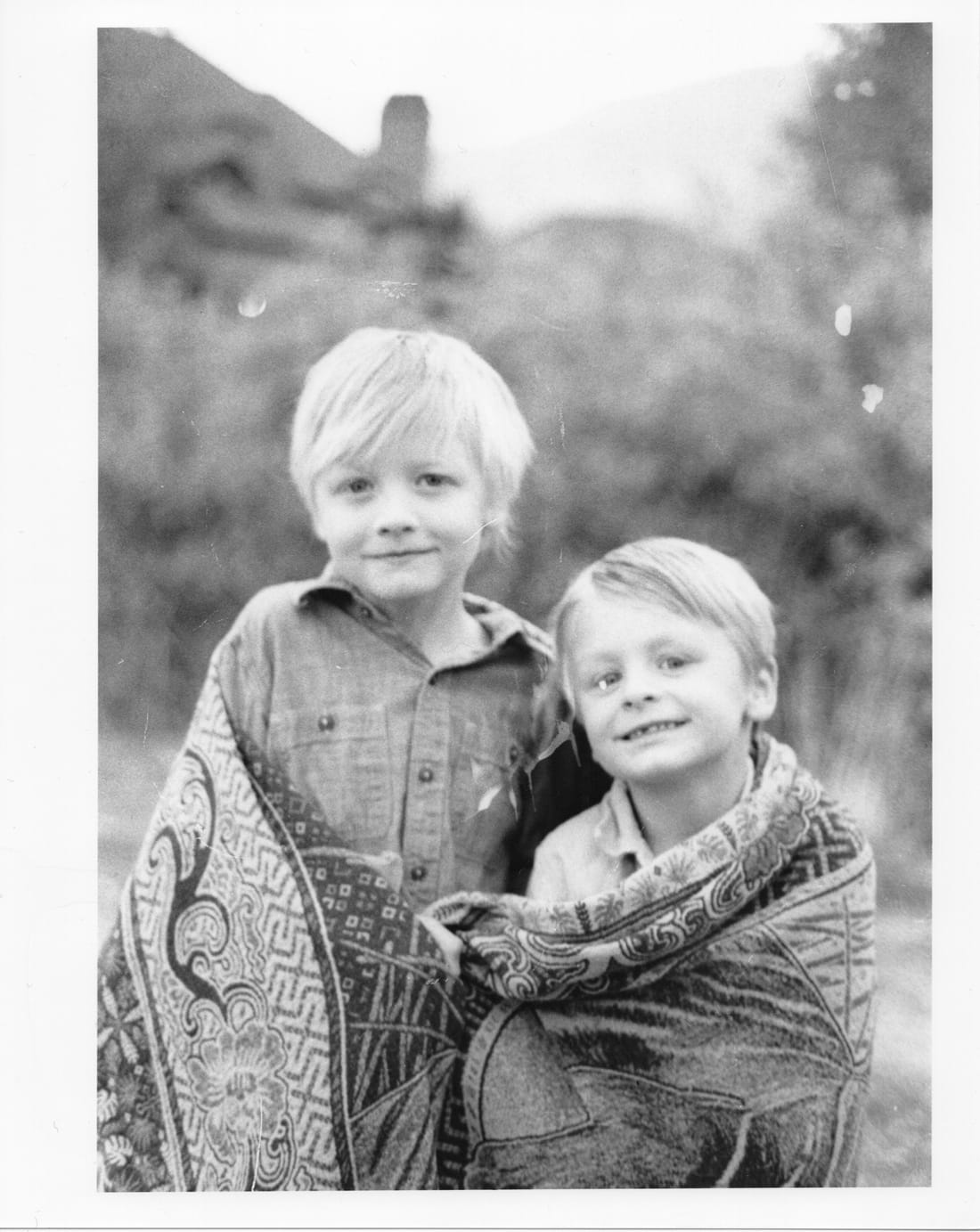Print of two smiling blond boys wrapped together in a patterned blanket.