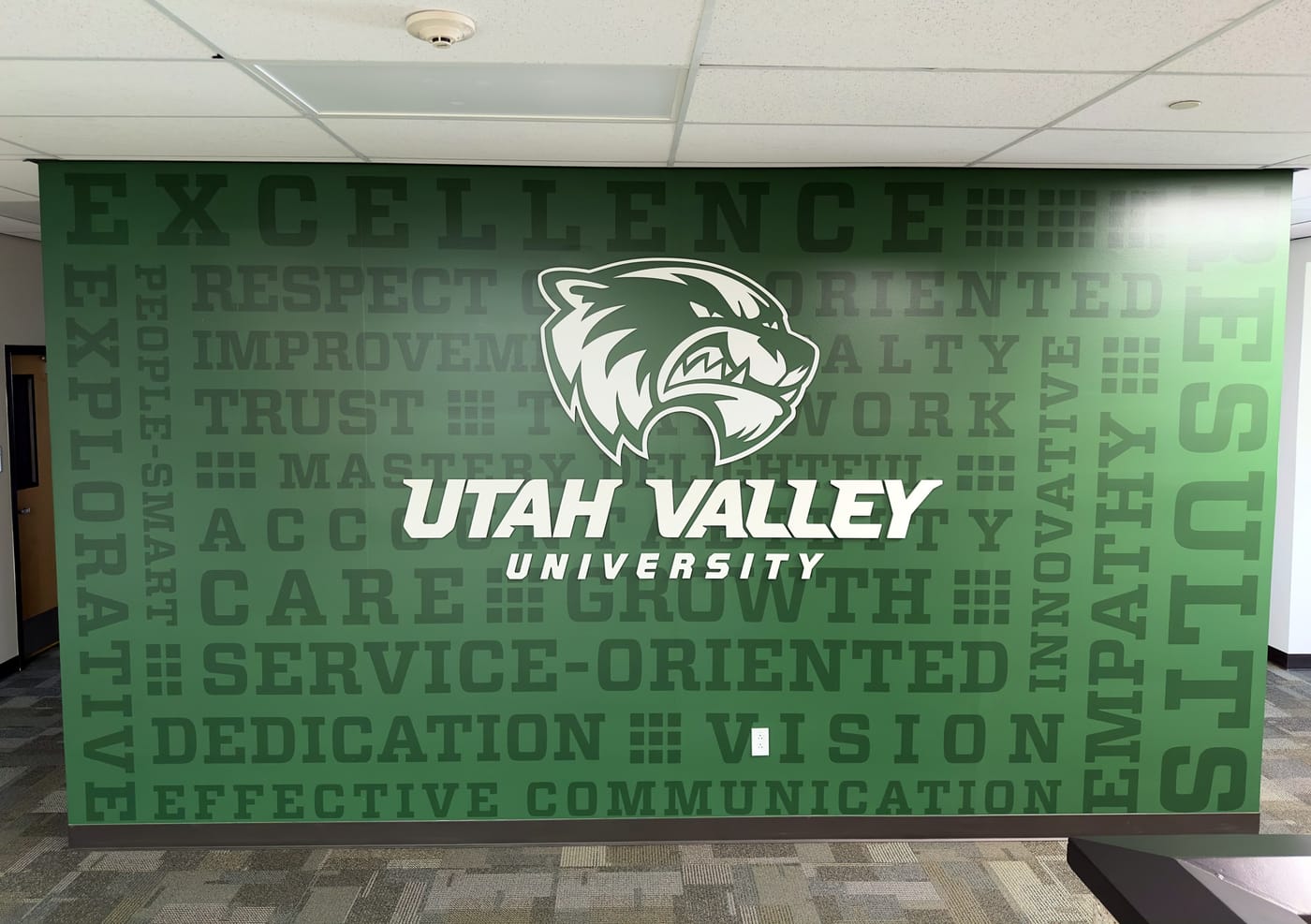 A green wall of stacked values words such as Excellence, Trust and Service-Oriented behind a large Utah Valley University wolverine logo.