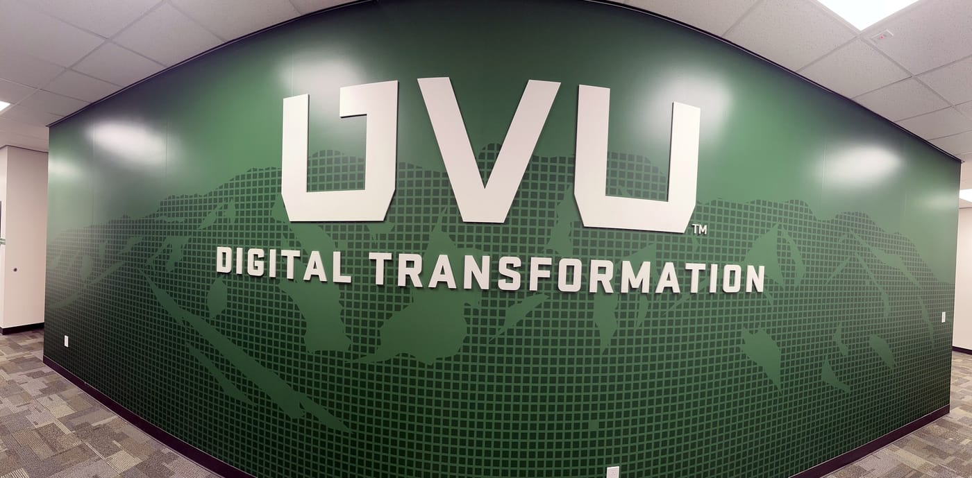 A curved hallway wall with large white UVU letters and the words Digital Transformation over a green pixel-grid mountain graphic.