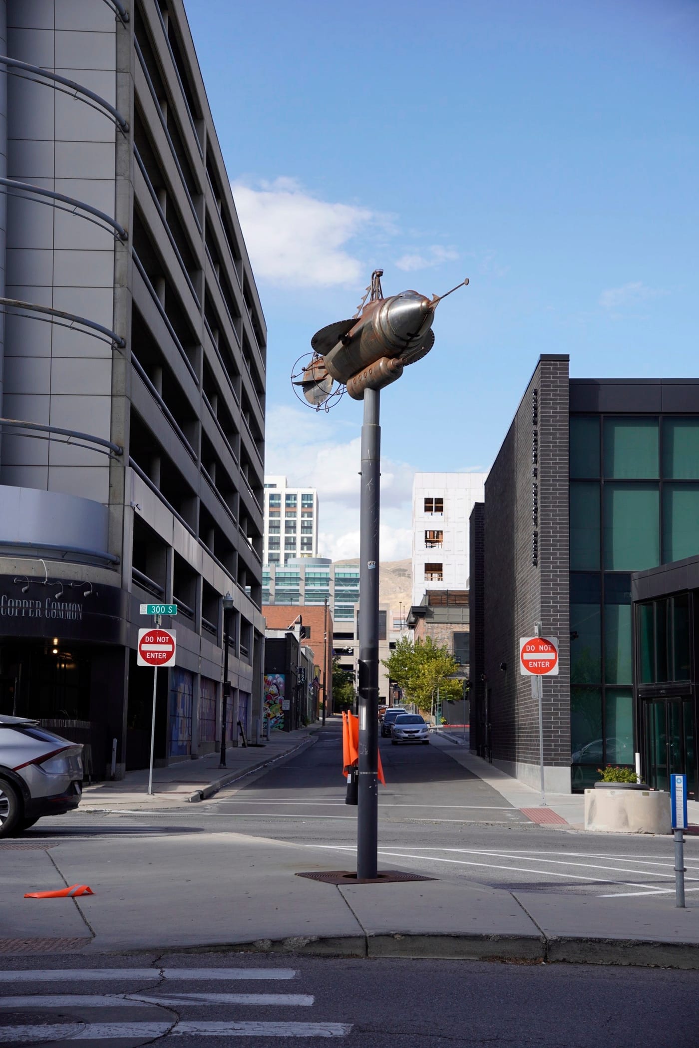 A rocket-shaped sculpture on a tall pole at a downtown intersection, between a parking structure and a glass office building.