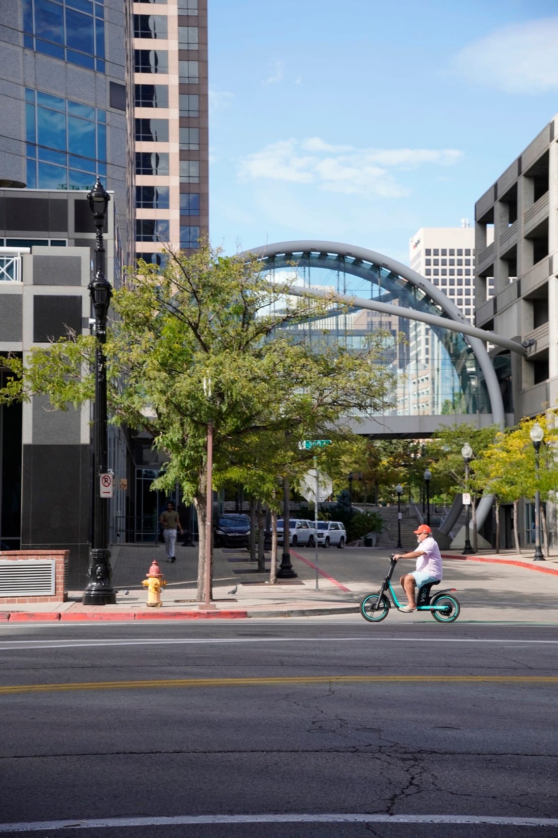 A rider on an e-scooter passes trees and a steel-arched glass skybridge between downtown towers.