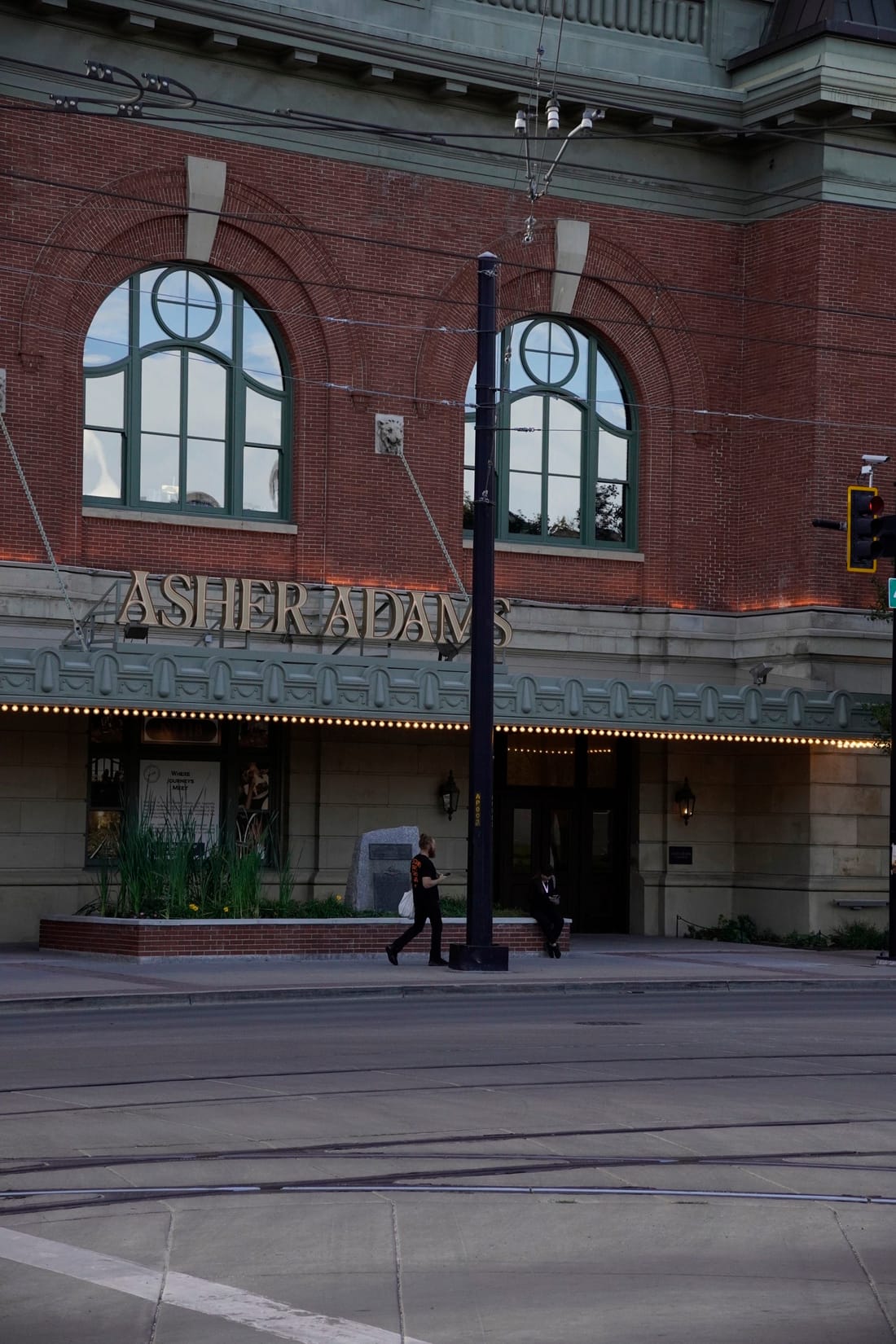A person walks past the Asher Adams hotel’s brick facade with arched windows and a lit marquee at dusk.