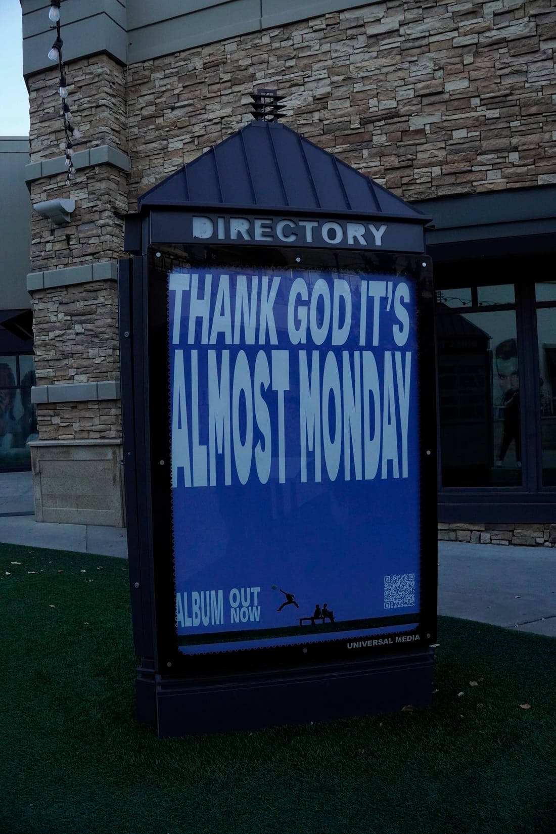 A lit directory kiosk at dusk showing a blue poster that reads “Thank God It’s Almost Monday.”