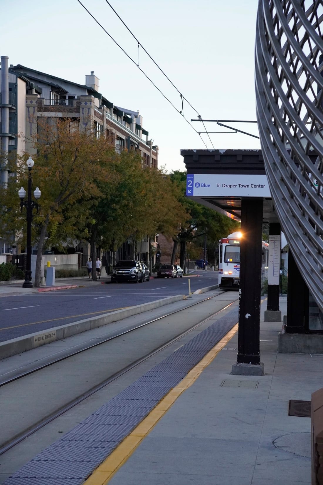A TRAX train approaches a platform at dusk, its headlight on, beside a curved metal sculpture.