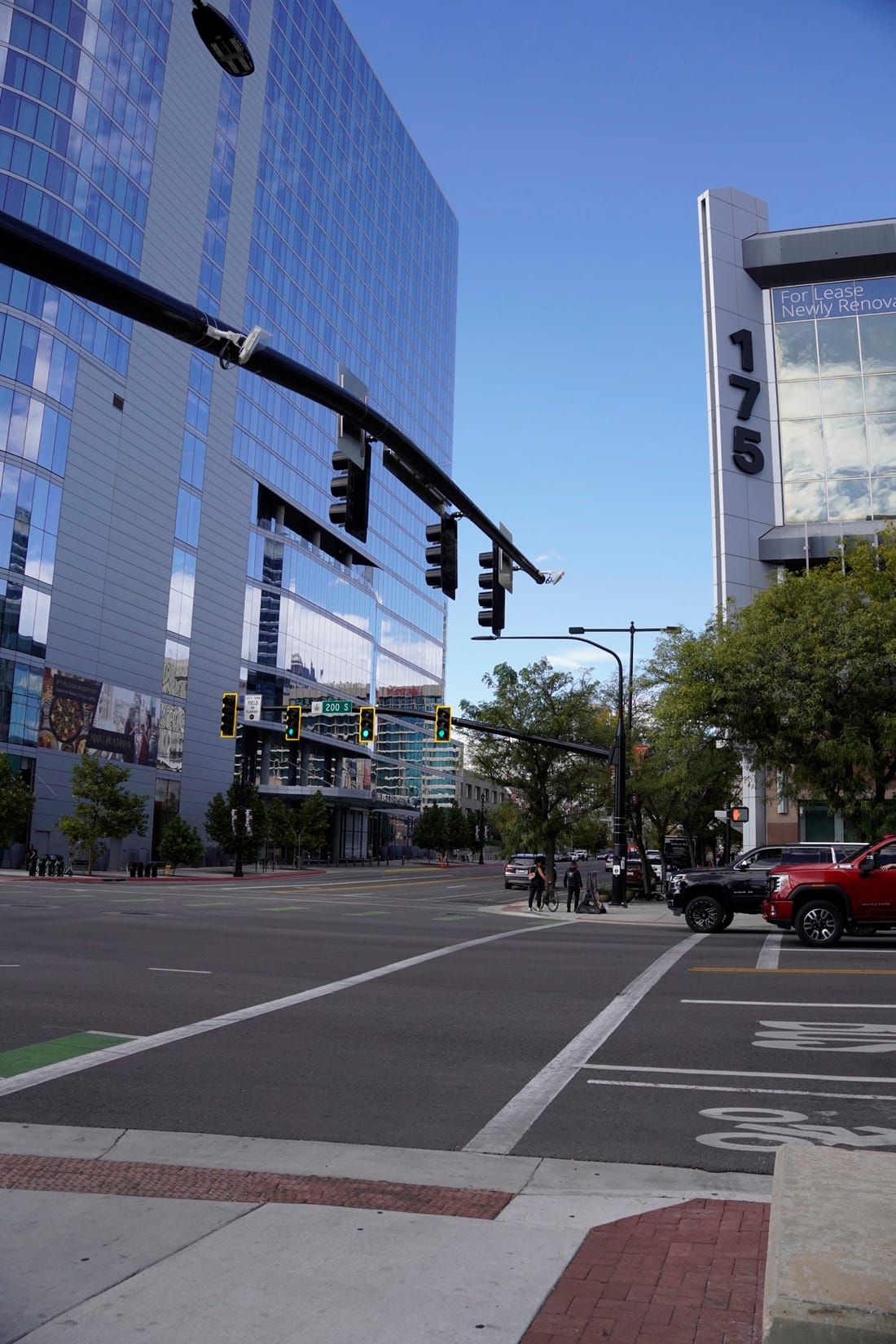 A wide downtown crosswalk under traffic signals, with glass towers and a building marked 175 on the right.