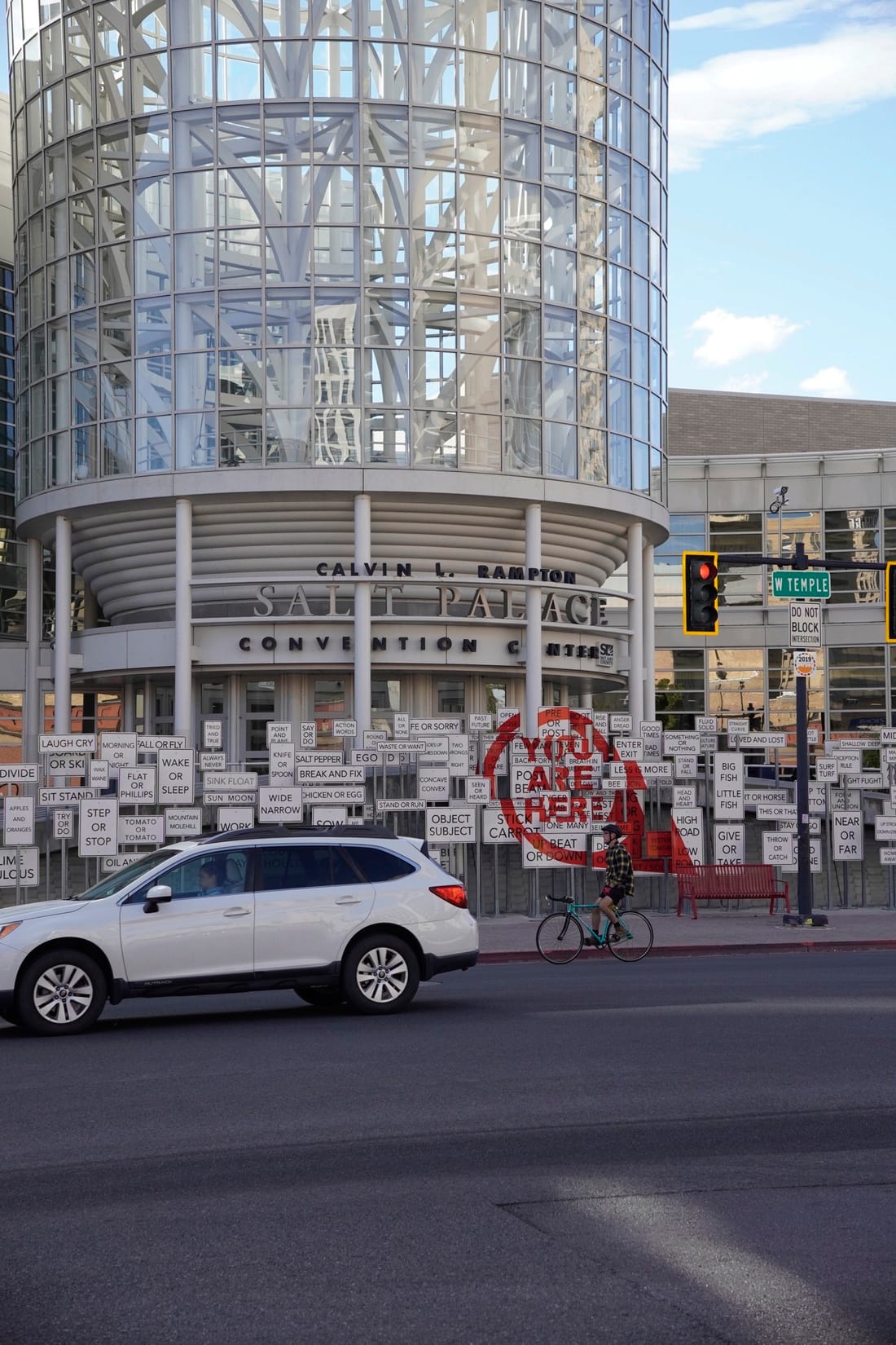 The round glass entrance of the Salt Palace Convention Center, with a fence of word signs and a red “You Are Here” sign.