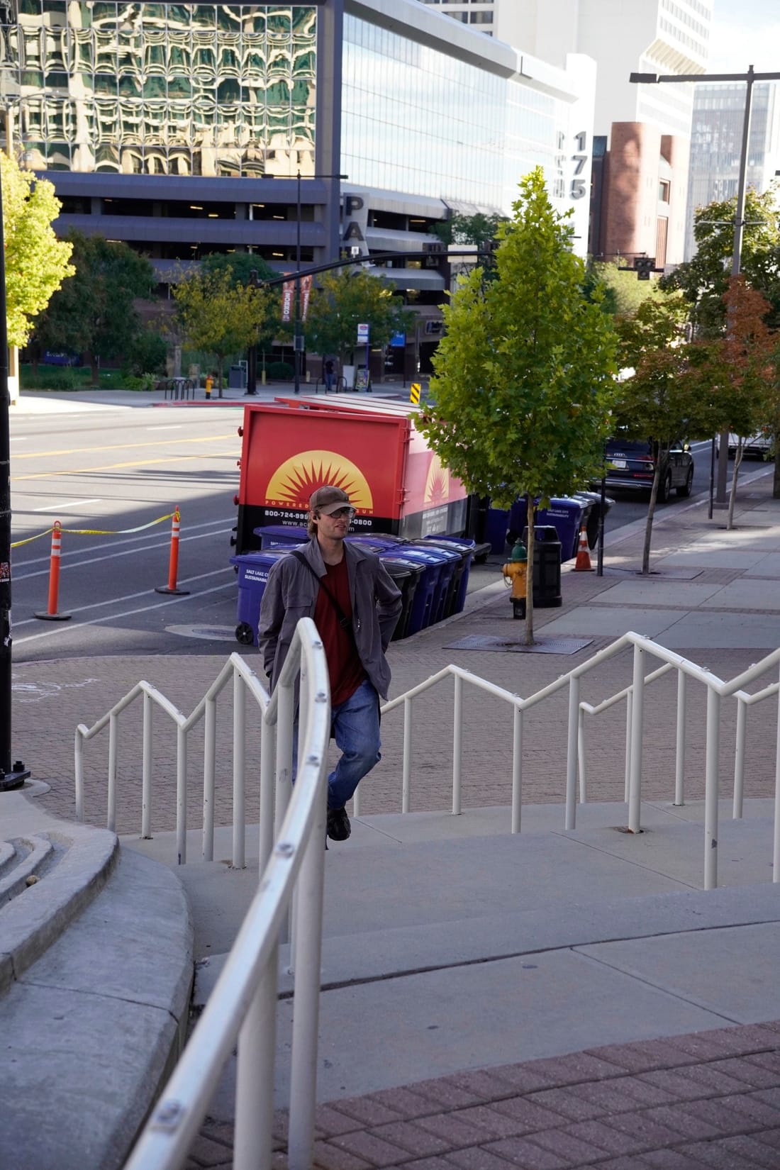 A man in a cap leans on a white railing at the top of outdoor steps, with a food truck and tall buildings behind him.