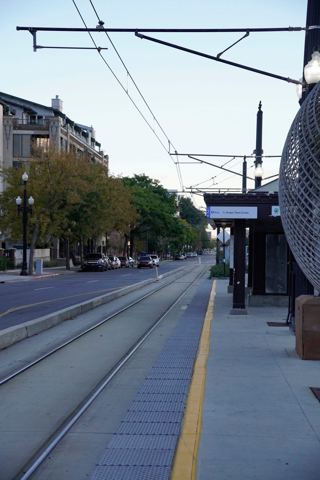 An empty TRAX platform at dusk, with rail tracks and overhead wires running toward tree-lined streets.