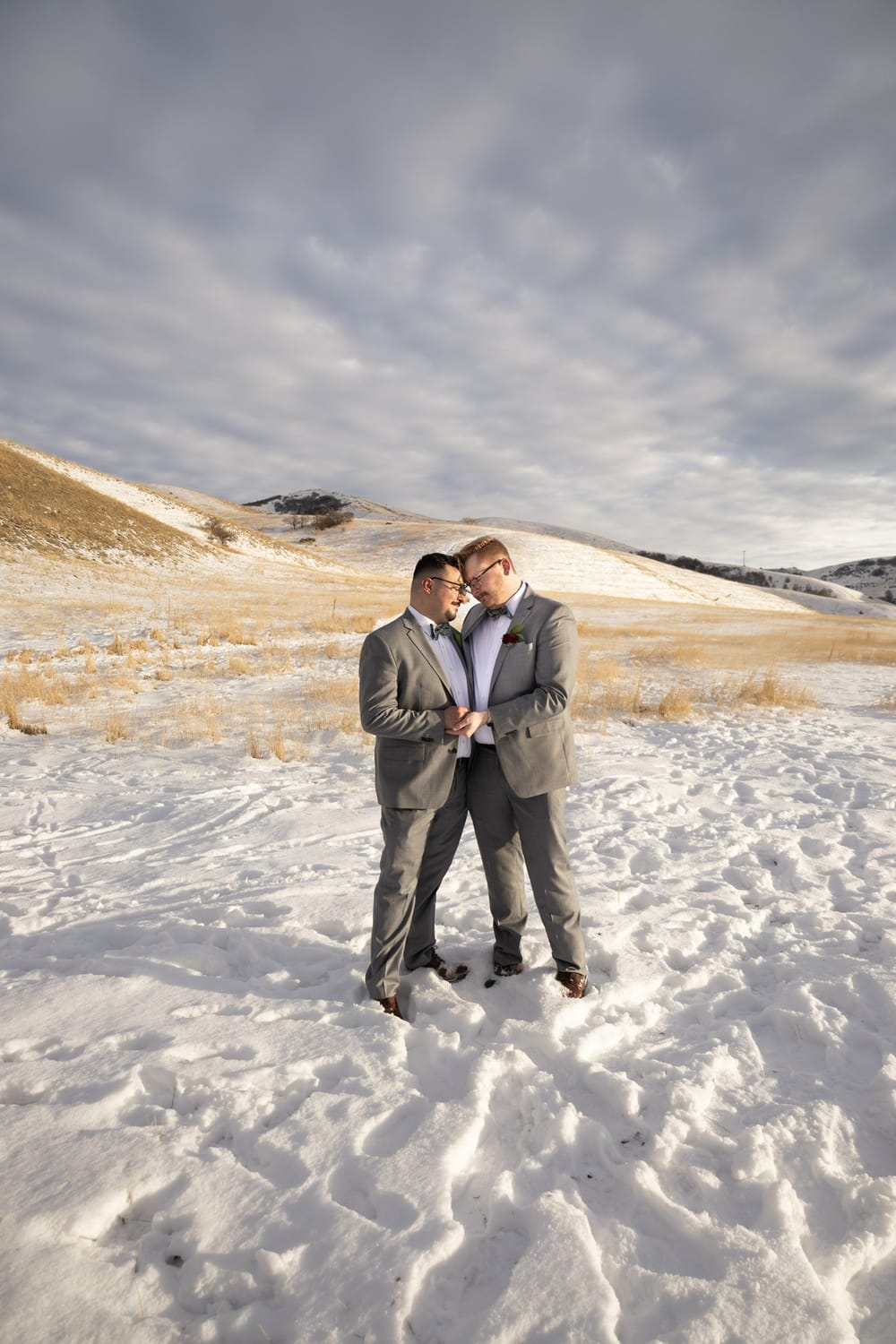 Paul and his husband Ely in grey suits, foreheads together, in snowy foothills.