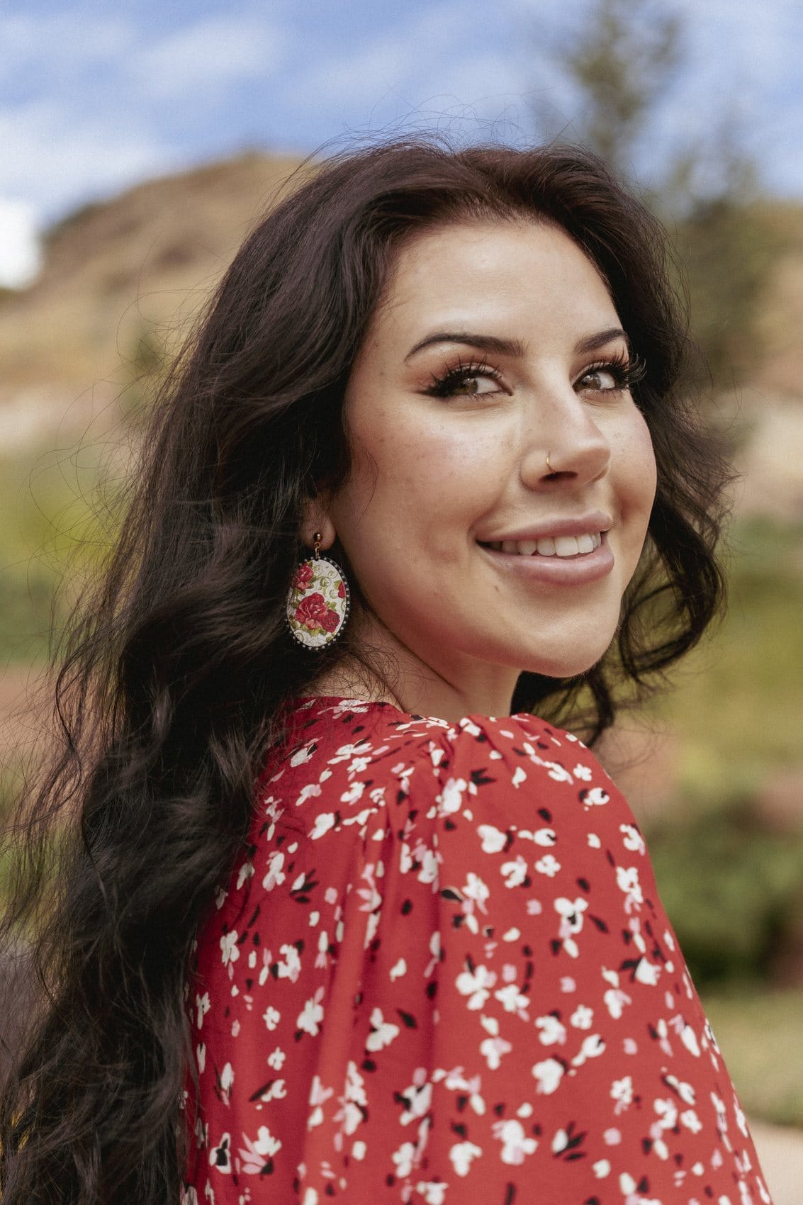 Outdoor portrait of a smiling woman with long dark hair and a red floral blouse, looking back over her shoulder against hills and sky.