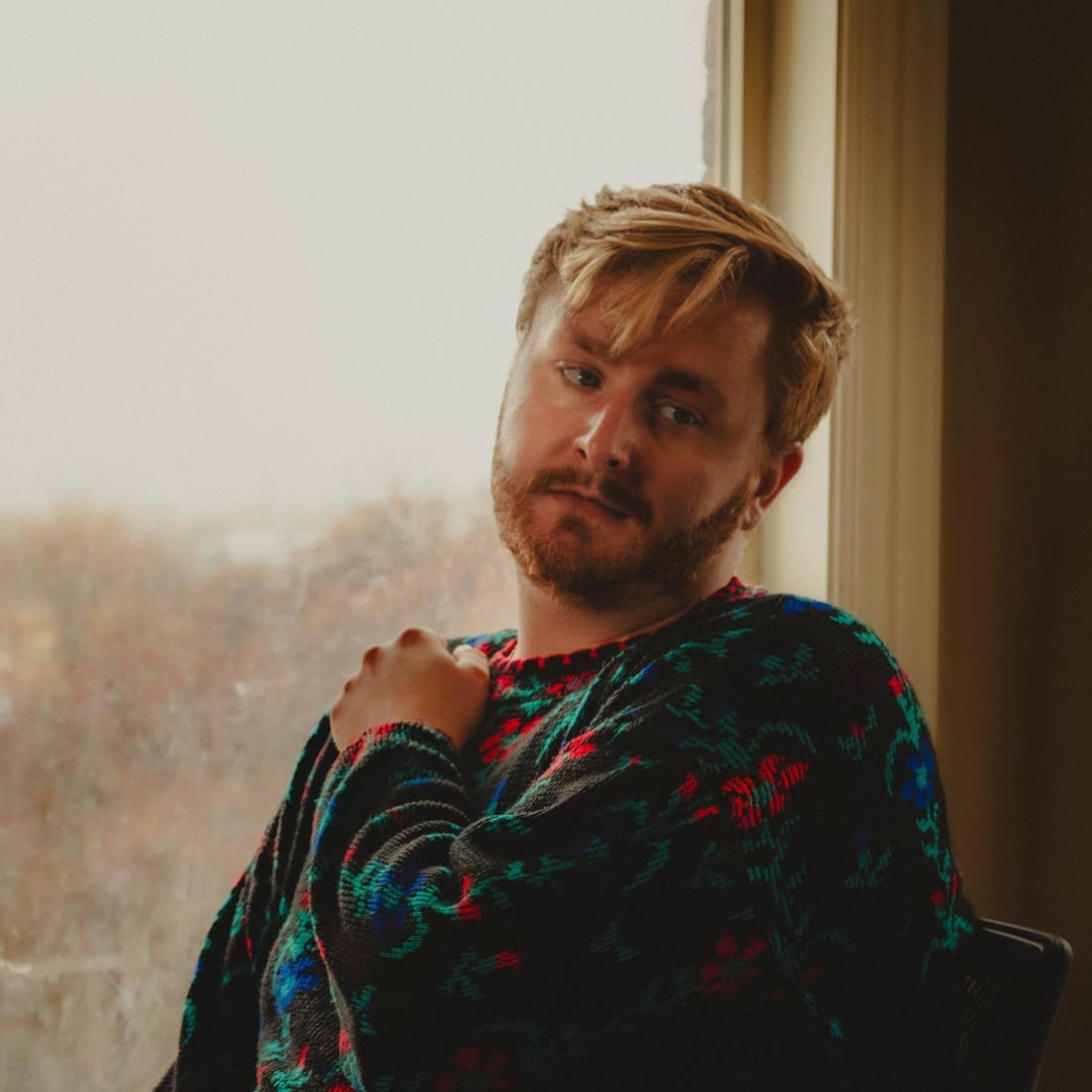 A bearded man in a patterned knit sweater sitting by a window in soft overcast light.