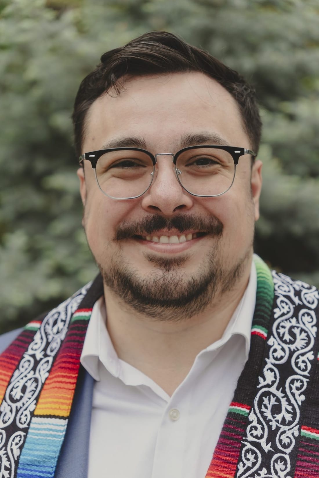 A smiling bearded man in glasses wearing a striped serape graduation stole, in front of evergreen foliage.