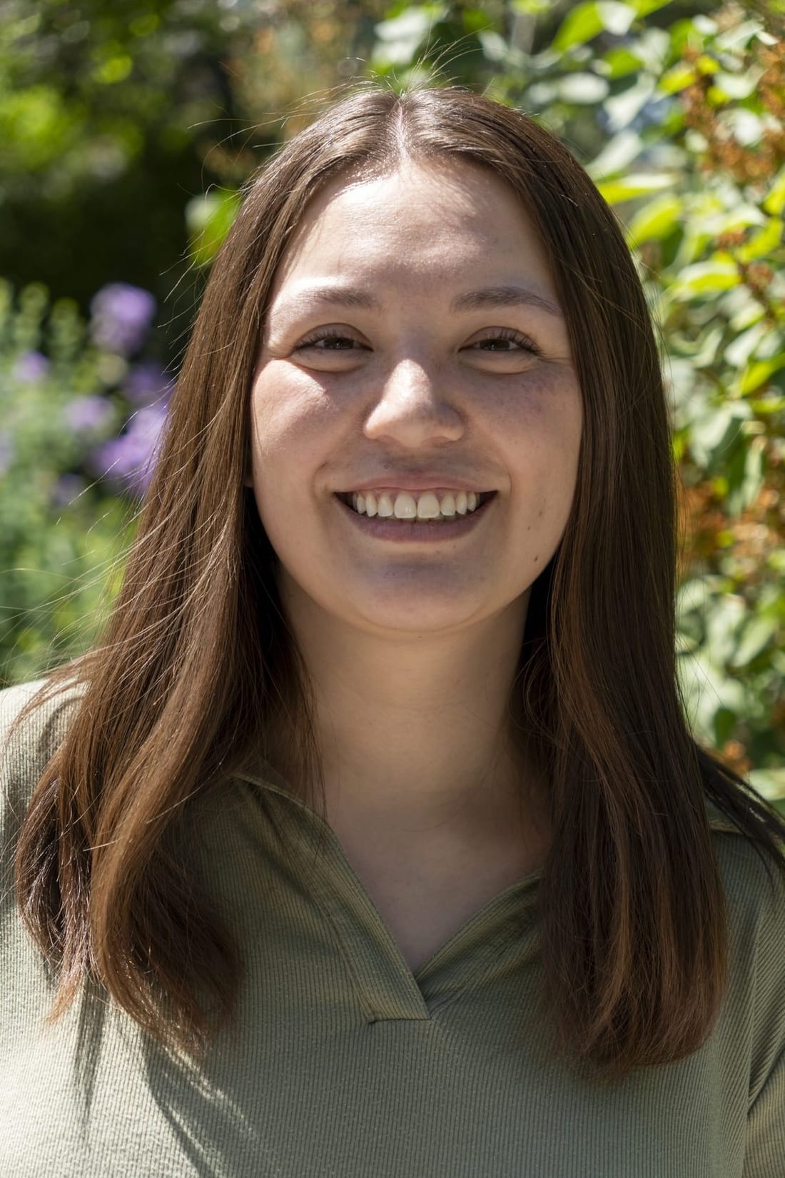 A smiling young woman with long brown hair and a sage collared top, in front of leafy shrubs and purple flowers.