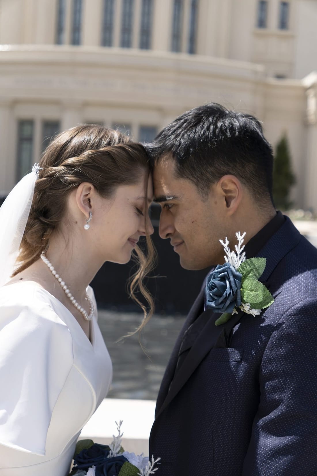 A bride and groom touching foreheads with eyes closed, in front of a white temple.