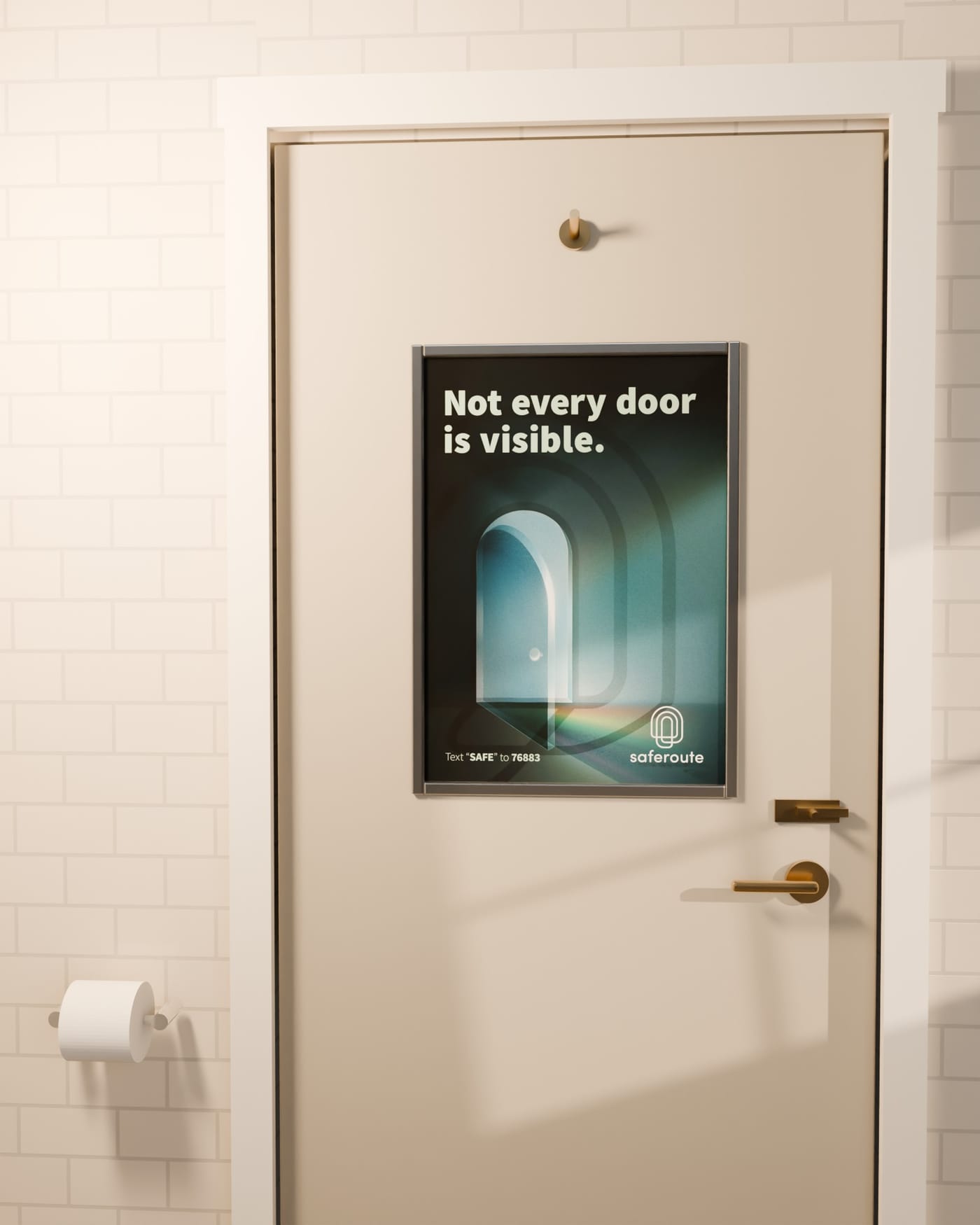 The SafeRoute poster, “Not every door is visible,” in a thin aluminum frame on the inside of a restroom door, with cream subway tile, a toilet-paper holder and warm late light.