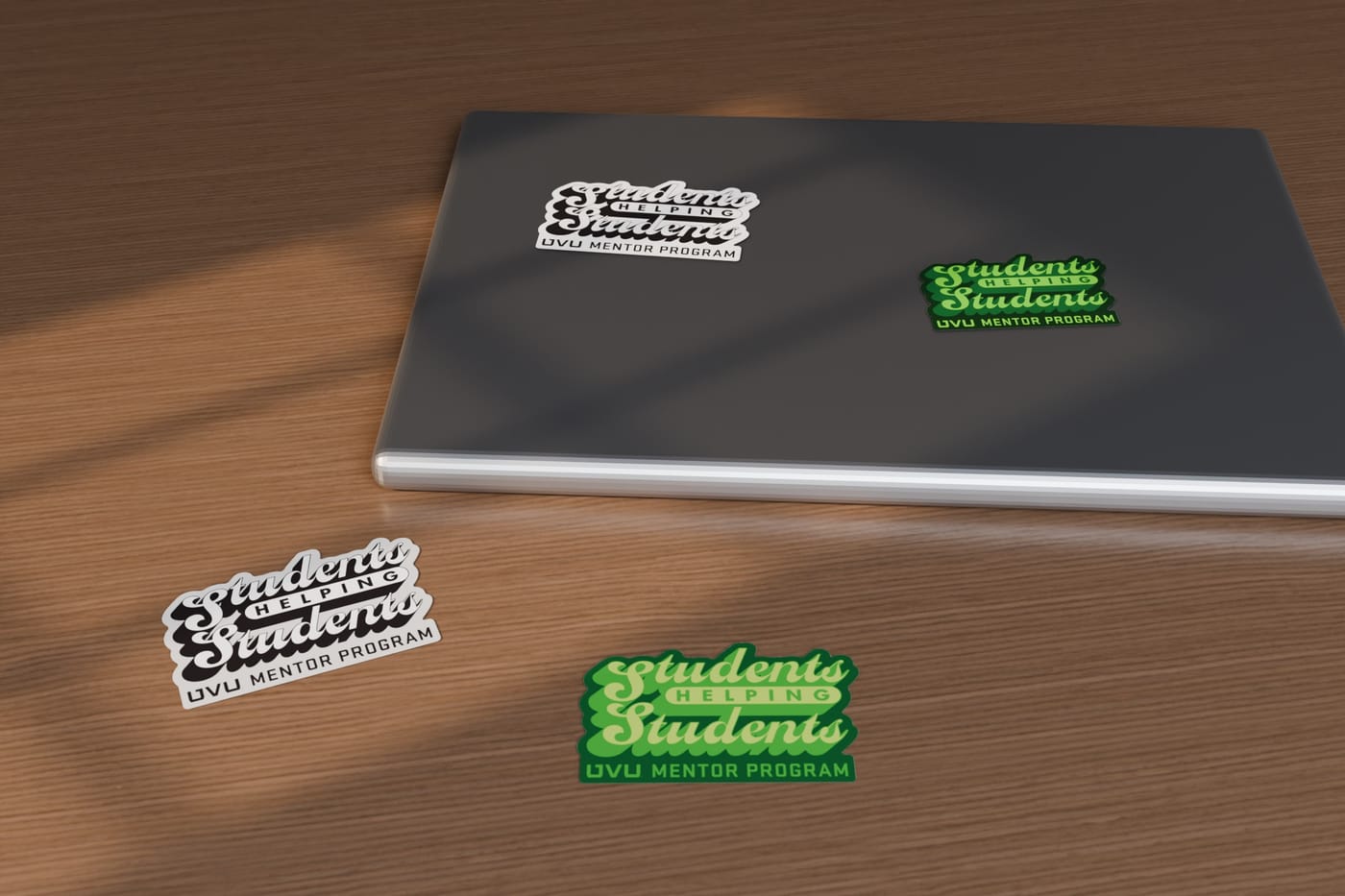 Students Helping Students die-cut stickers in black-and-white and green, on a laptop lid and a wooden desk in window light.