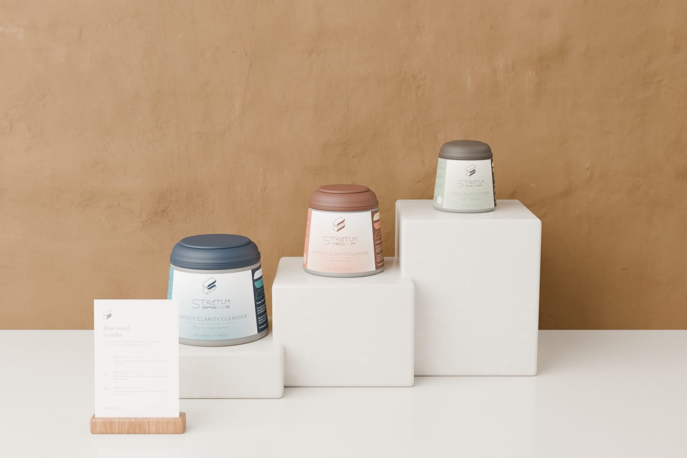 A retail display of three stepped stone tiers, one Stratum jar per tier rising from BASIS on the lowest to PRIMA on the highest, with a small printed card in an oak stand in front.