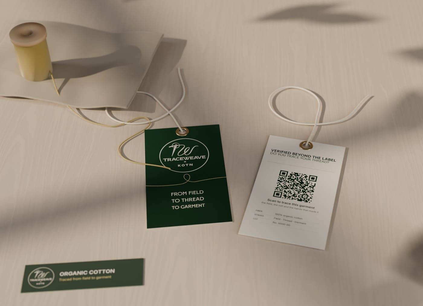 Two TraceWeave × KOTN hang tags on cotton twine, the green front and the cream back with a QR code, beside a woven ORGANIC COTTON label, a spool of pale-gold thread, a cotton boll and a fold of undyed cotton, in leafy window light.